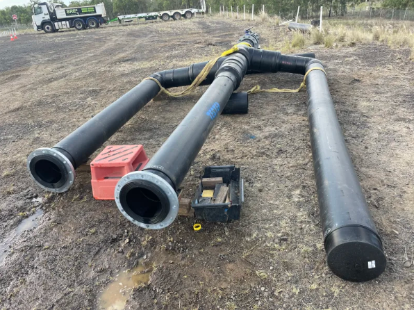 Large black industrial pipes with metal flanges are laid out on a dirt ground, connected in a T-shape. Nearby are a red crate, a black toolbox, and some small equipment. A truck is visible in the background.