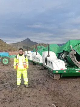 Person in a bright yellow high-visibility suit stands on a muddy construction site, with large covered machinery and distant hills under a cloudy sky in the background.