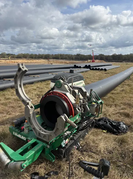 A large pipe fusion machine is set up in a grassy field, welding sections of black plastic pipe. More pipes are laid out in the background, with construction equipment visible under a cloudy sky.