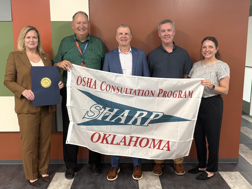 Five people stand inside holding a banner that reads OSHA Consultation Program SHARP Oklahoma. One person on the left holds a folder with a gold seal. They are smiling and standing against a multicolored wall.