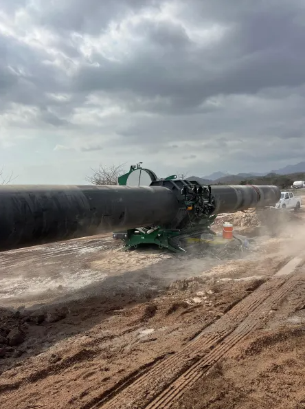 A large pipeline is being welded together on a dirt construction site under a cloudy sky, with machinery holding the pipes in place and vehicles in the background.