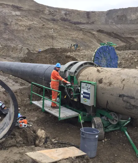A worker in orange coveralls and a blue helmet operates a large pipe fusion machine, joining sections of pipeline at a construction site with dirt and rocky terrain in the background.
