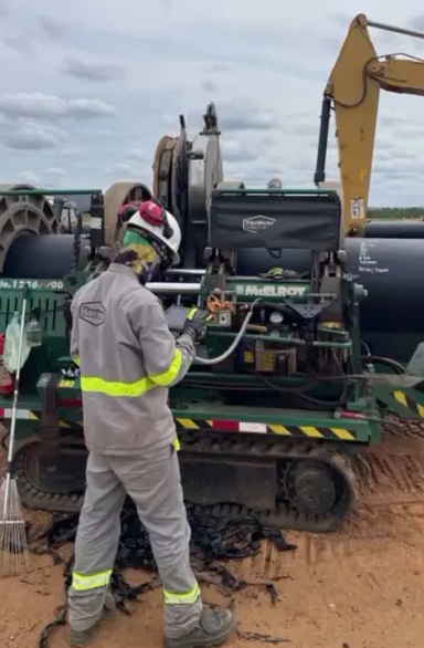 A construction worker in safety gear operates a large green pipe fusion machine on a sandy worksite, with heavy equipment and pipes visible in the background.