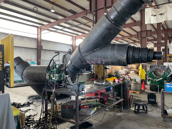 Large black industrial pipes are being welded together inside a spacious warehouse filled with machinery, tools, and equipment. The workspace is cluttered, with cables, stools, and materials scattered around.