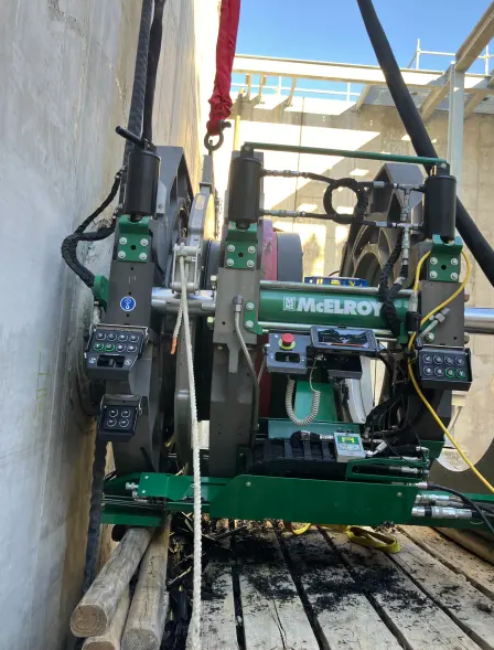 A McElroy pipe fusion machine is positioned vertically on a construction site, attached to large pipes with control panels and cables, on a wooden platform against a concrete wall.