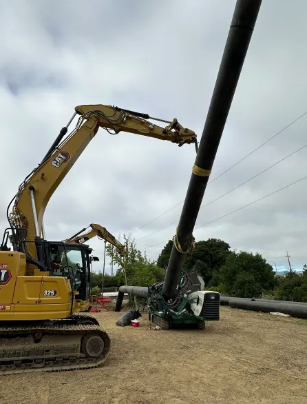 A yellow excavator lifts a large black pipe vertically using straps at a construction site with workers nearby, under a cloudy sky. More equipment and pipes are visible in the background.