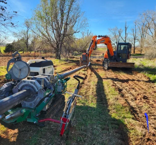 An excavator positions a long pipe in a dirt trench while a pipe fusion machine is set up on the ground. Trees and grass surround the rural construction site under a clear blue sky.