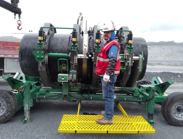 A worker in a red safety vest and hard hat stands on a yellow platform next to a large green industrial pipe fusion machine on a construction site with rocky terrain in the background.