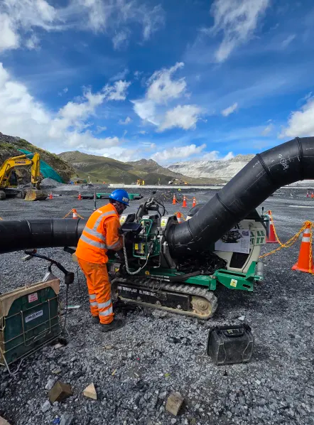 A construction worker in orange safety gear operates heavy machinery to join large black pipes at an outdoor worksite, with mountains and construction equipment visible in the background.