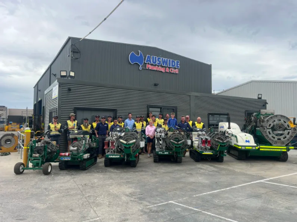 A group of people in high-visibility clothing stand in front of green industrial machinery and a building with an AUSWIDE Plumbing & Civil sign on it, under a cloudy sky.