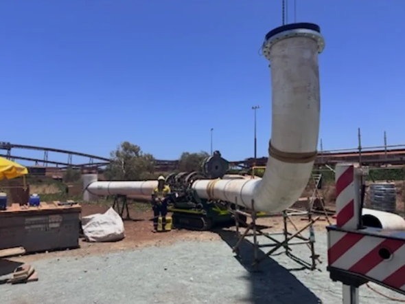 A large white industrial pipeline is being installed outdoors, supported by scaffolding. Two workers in safety gear operate equipment under the pipe. The sky is clear and blue, with structures visible in the background.