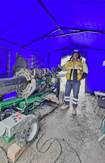 A construction worker in safety gear stands next to a large pipe fusion machine inside a blue tent at a worksite.