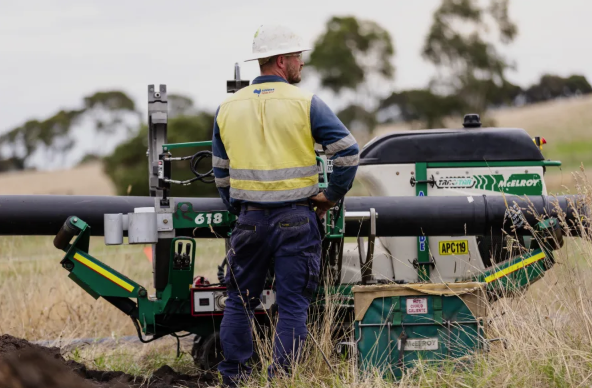 A construction worker in a hard hat and high-visibility vest stands next to a green pipe fusion machine in a grassy outdoor area with trees in the background.