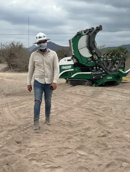 A construction worker wearing a hard hat, mask, and jeans walks on a sandy site with a large green industrial machine in the background under a cloudy sky.