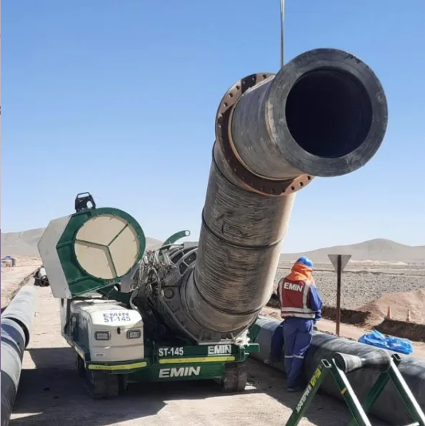 A large industrial pipe is being installed by workers in safety gear on a construction site in a desert area, with machinery supporting the pipe and a clear blue sky overhead.