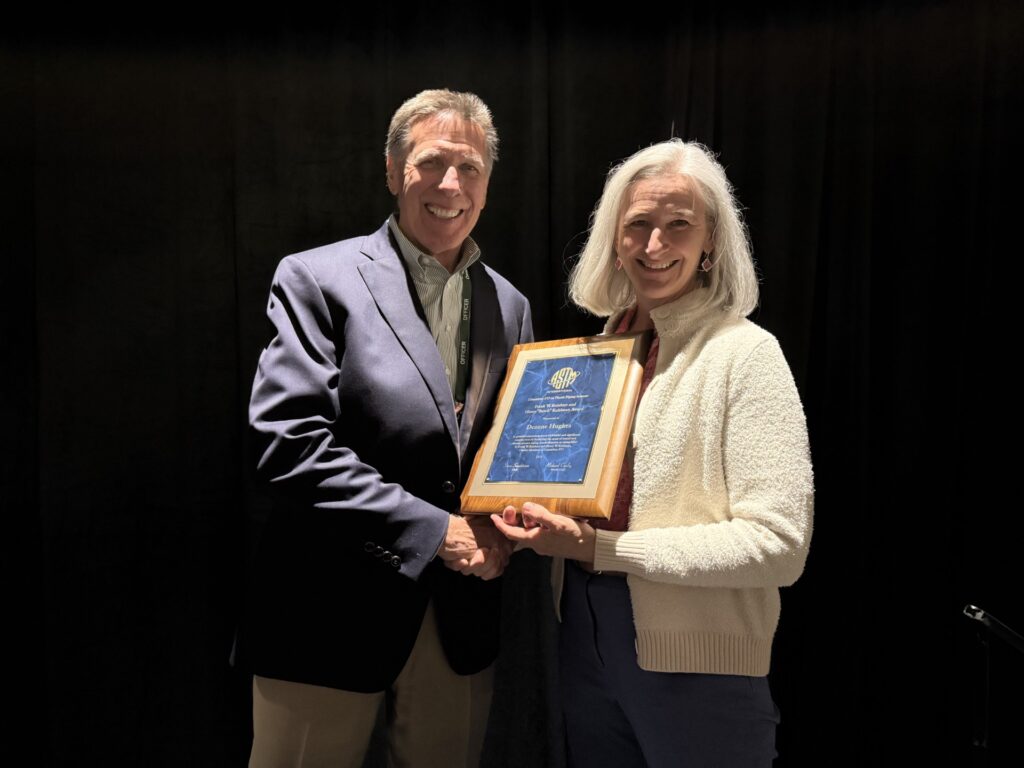 Two people stand in front of a dark background, smiling. The person on the right holds a blue and gold plaque while both shake hands, suggesting an award presentation or recognition event.