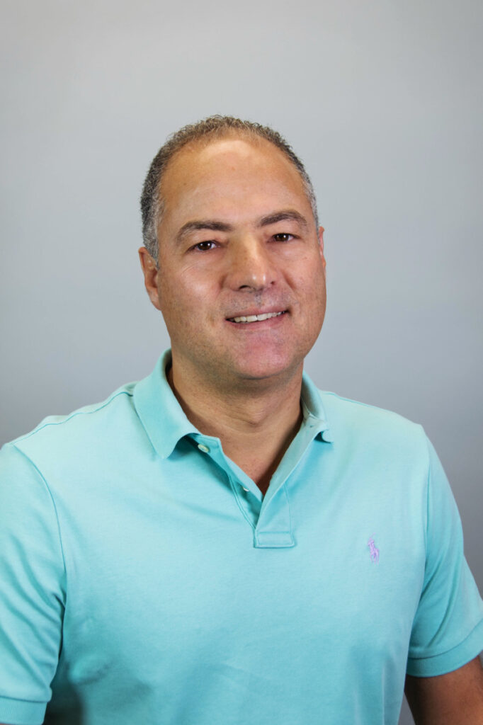 A man with short, graying hair smiles at the camera. He is wearing a light blue polo shirt and is posed in front of a plain, light gray background.
