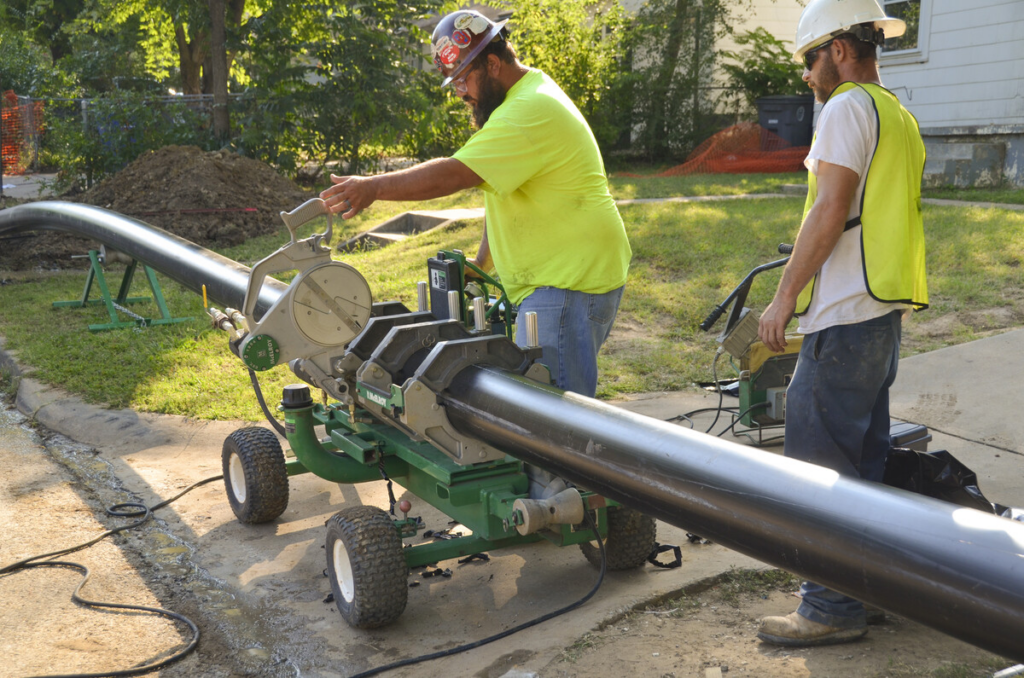 Two construction workers in safety gear operate a machine to fuse sections of large black plastic pipe outdoors on a residential street. One worker adjusts the machine while the other monitors the process.
