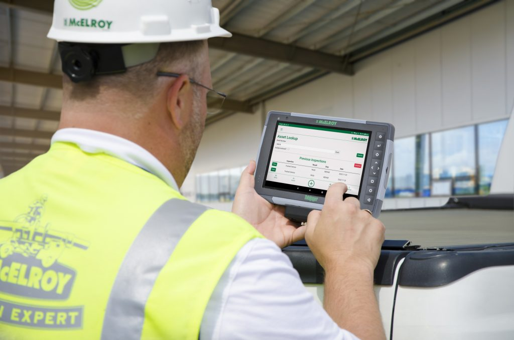 A construction worker wearing a hard hat and safety vest uses a tablet mounted on a vehicle, interacting with a digital interface on the screen at an outdoor industrial site.