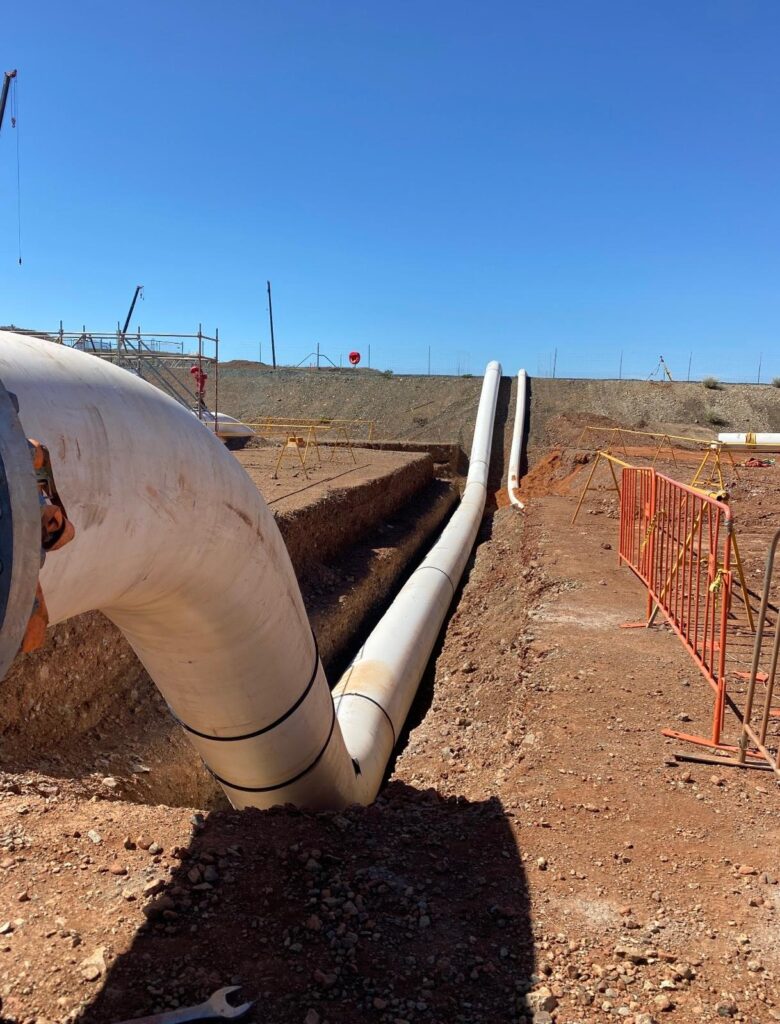 Large white pipelines run through a dirt trench at a construction site under a clear blue sky, with barriers and equipment visible in the background.