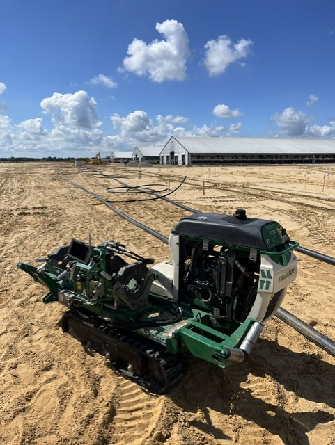 A small green machine sits on sandy ground near long pipes, with large barns or industrial buildings and a blue sky with clouds in the background.