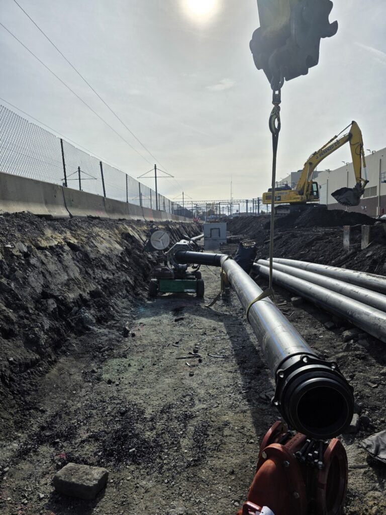 A construction site with large metal pipes, a crane lifting a pipe, and an excavator in the background, all under a bright sun and clear sky.