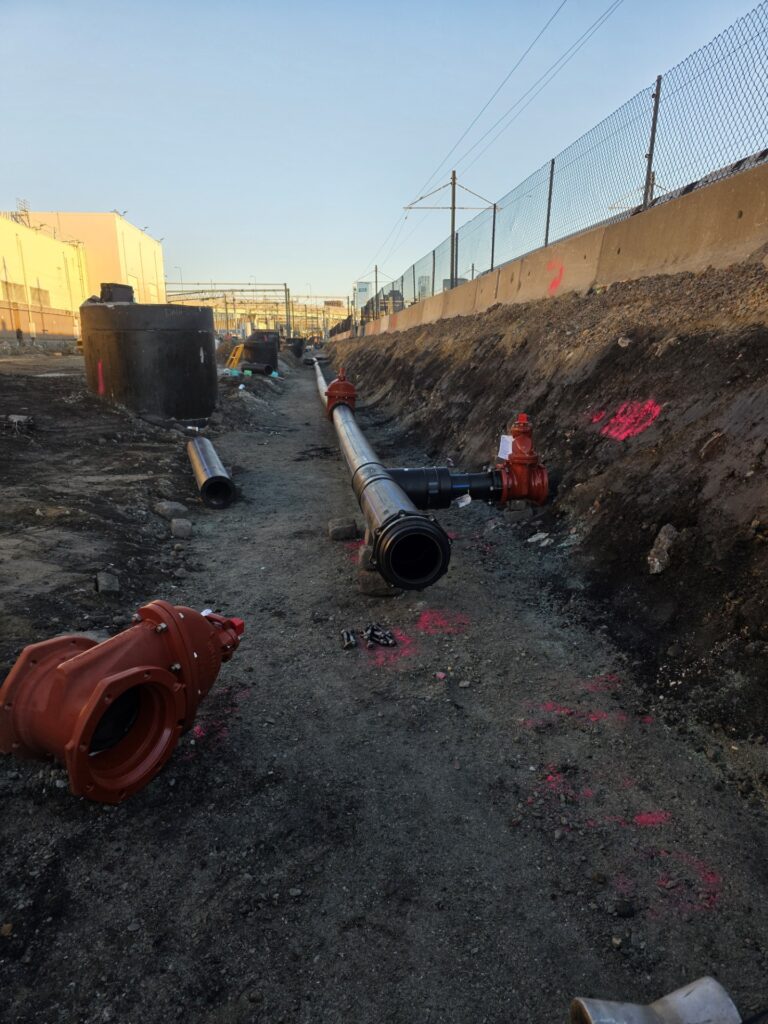 A construction site with large pipes and fittings laid out in a trench, surrounded by dirt and concrete barriers, with buildings and a fence in the background.