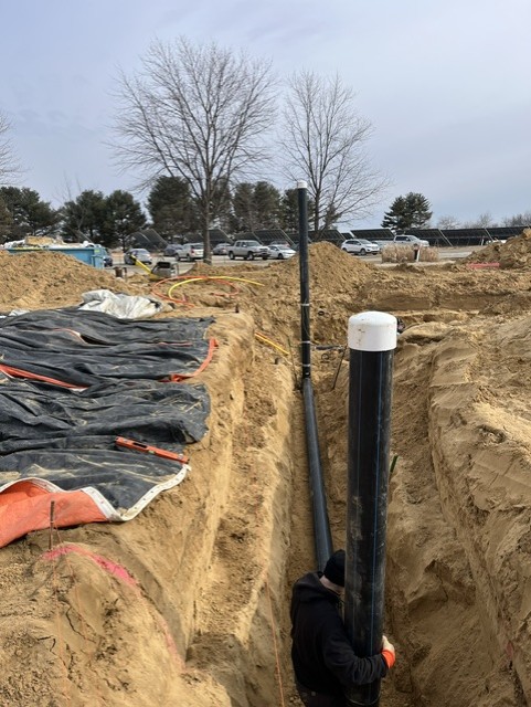 A worker in a trench installs large black pipes in a sandy construction site, with trees and parked cars visible in the background.