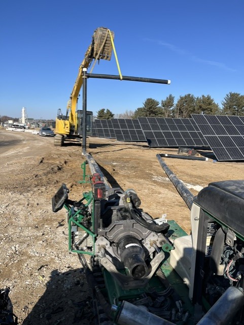 A yellow excavator lifts a long pipe near rows of solar panels at a construction site under a clear blue sky. Dirt and machinery are visible in the foreground.