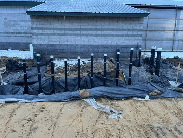 Several black pipes with white caps are set vertically in the ground in front of a building with gray bricks. The area is covered with black plastic and surrounded by dirt and sand.