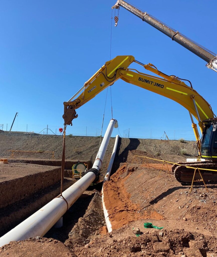 A yellow excavator lifts a large white pipe at a construction site, with another crane and pipes in the background on a sunny day.