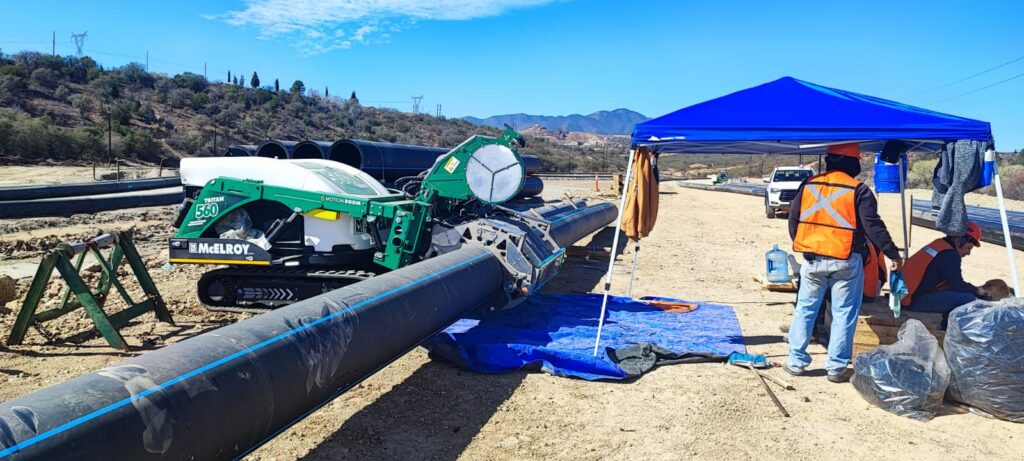 Workers in safety vests operate heavy machinery to weld large black pipes under a blue canopy at a construction site, with hills and power lines visible in the background.
