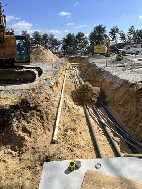 A construction site with a long trench containing several black pipes and a yellow pipe. Excavators and vehicles are in the background, and piles of dirt are alongside the trench under a clear blue sky.
