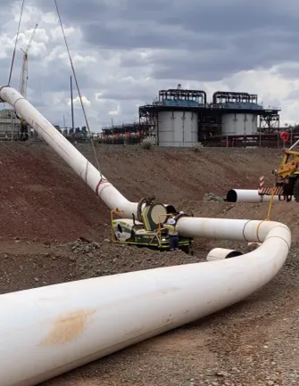 Large white pipes being installed at an industrial site, with workers and machinery present; two large storage tanks and cloudy skies are seen in the background.