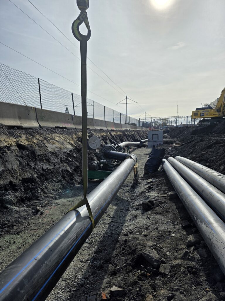 Large metal pipes being installed in a trench at a construction site, with machinery and a fence visible under a bright sky.