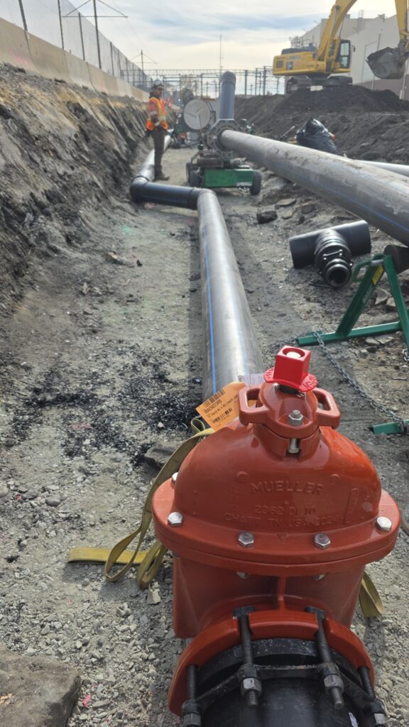 A construction site with large metal pipes being installed underground, a red valve in the foreground, and a worker wearing a safety vest and helmet standing in the background.