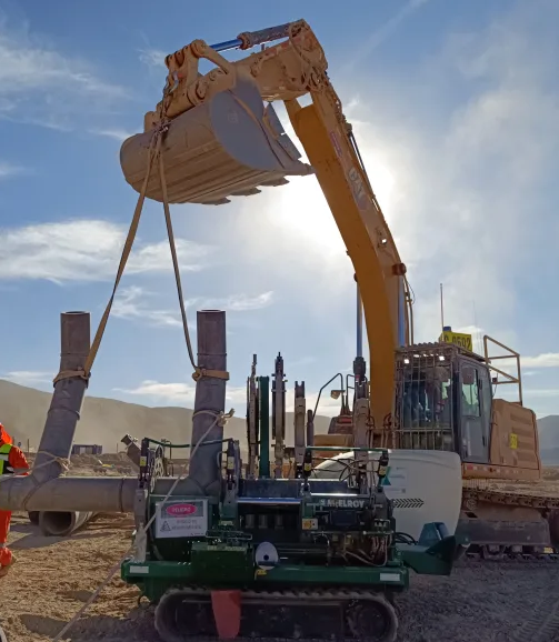 A large excavator lifts several pipes using straps at a dusty construction site, while a compact pipe fusion machine sits nearby. Sunlight shines behind the equipment, casting shadows and creating a bright, backlit scene.