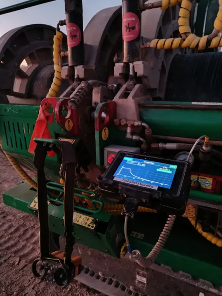 Close-up of agricultural machinery with yellow hoses, control valves, and a mounted tablet displaying a graph on the screen, likely monitoring equipment performance in a field setting.