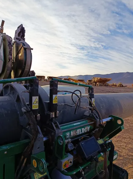 A close-up of a McElroy machine fusing a large pipe at a construction site, with dump trucks and mountains visible in the background under a partly cloudy sky.