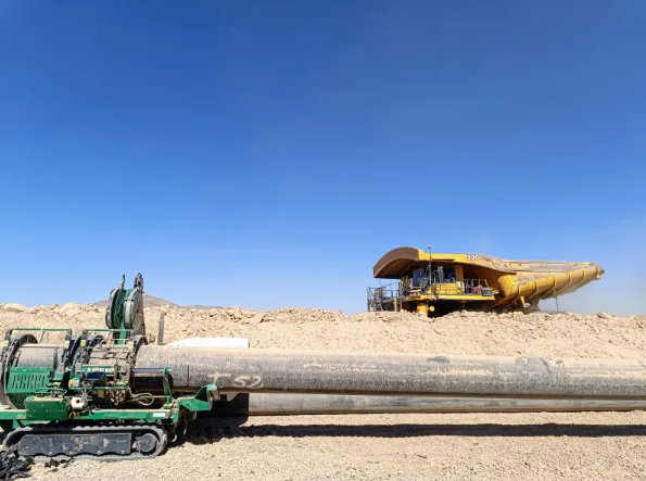 A construction site with heavy machinery: a green pipe layer on the left, a large yellow earthmover in the background, and a long pipe lying on the ground under a clear blue sky.