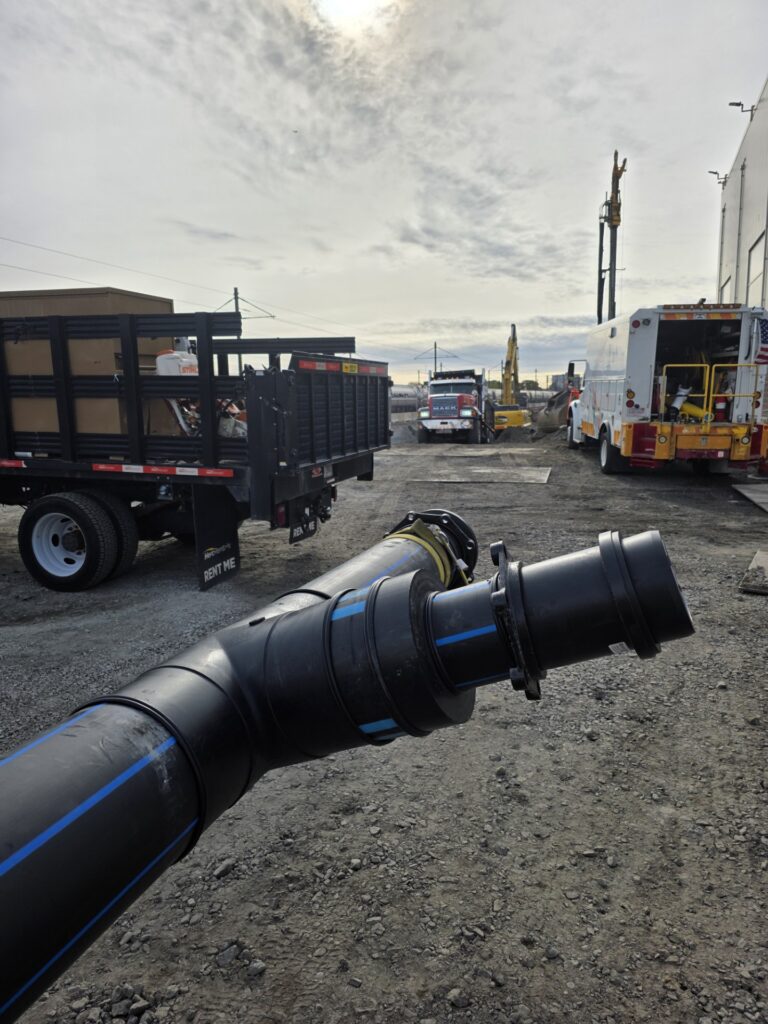 A large black pipe is held in the foreground at a construction site with trucks, trailers, equipment, and a cloudy sky in the background. The ground is gravel and dirt.