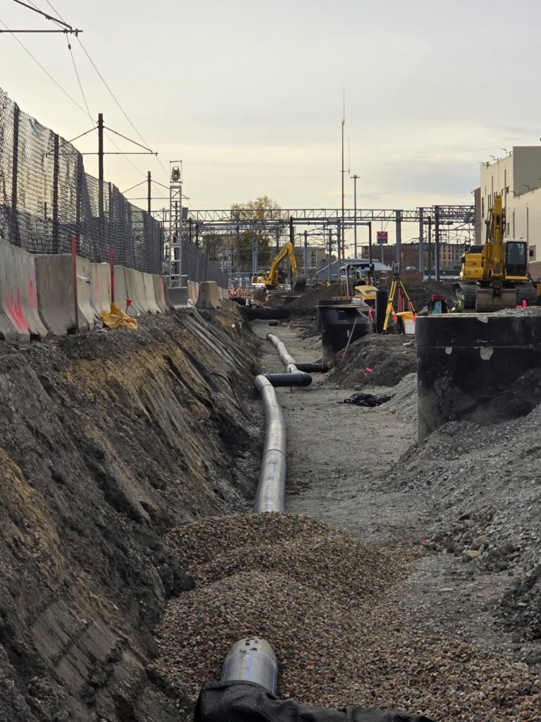 A construction site with a deep trench, exposed piping, gravel, heavy machinery, and workers in safety gear. Concrete barriers and fences line the sides; a railway track and industrial buildings are visible in the background.