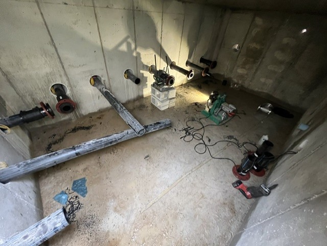 A concrete room with several pipes protruding from the walls, tools, and equipment on the floor, including a welding machine, cables, and pipe fittings scattered around.